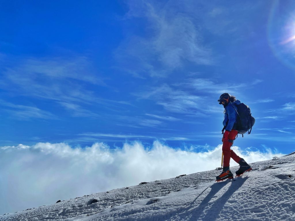 A man walks down a snowfield with crampons, a backpack, and many winter layers.