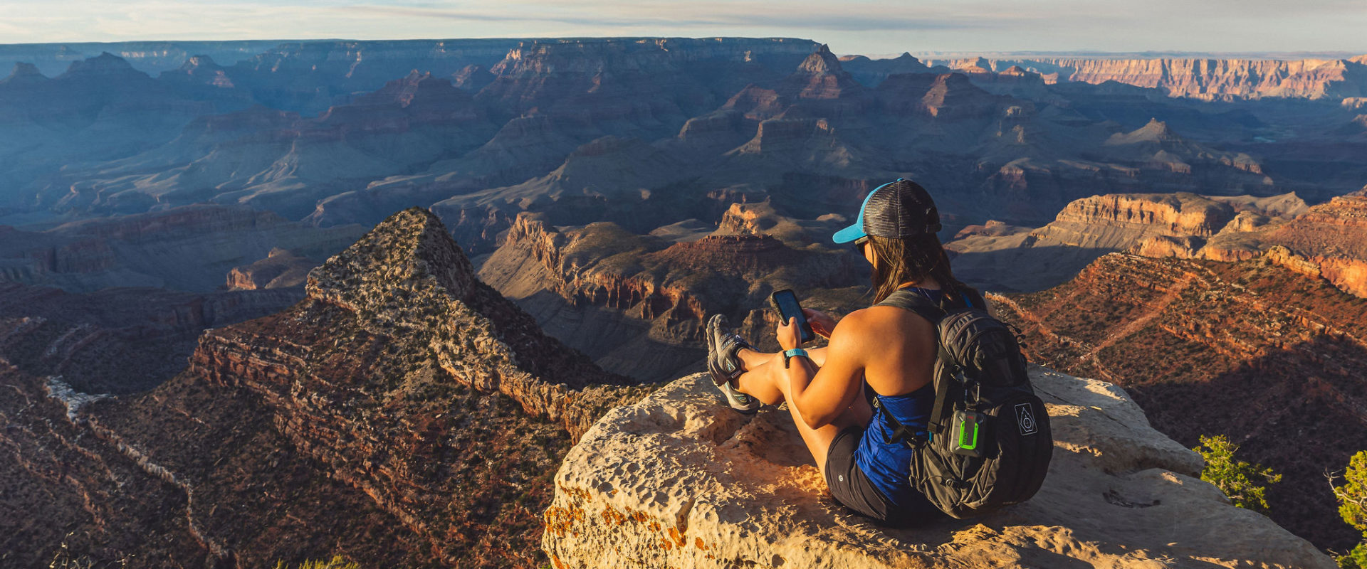 A person sits at the edge of a canyon holding her phone, with a ZOLEO unit attached to her backpack.