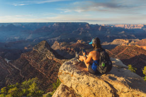 A person sits at the edge of a canyon holding her phone, with a ZOLEO unit attached to her backpack.