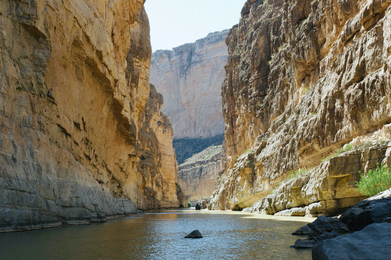 A desert canyon, with steep rock walls and a calm river flowing through the bottom of the canyon.  