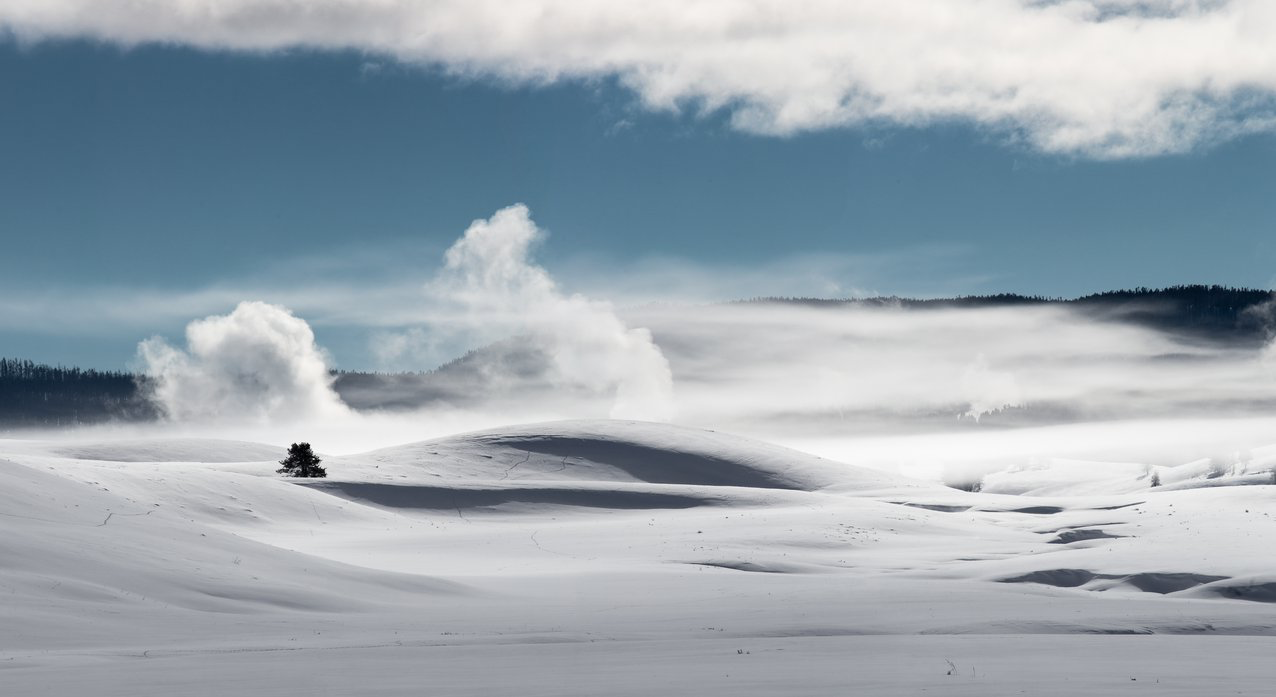 Wide open snow field with steam rising from geothermal pools, under a blue sky.