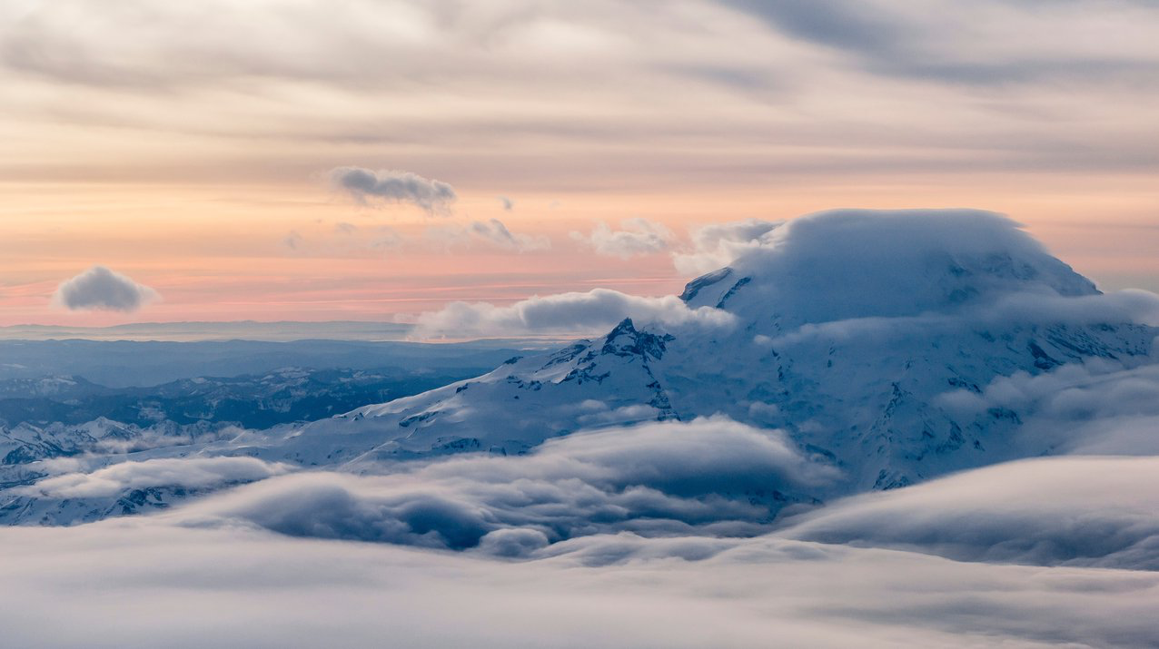 Snowy Mount Rainier pokes above the clouds with a colorful, pastel sunset of orange and soft pink hues int he background.
