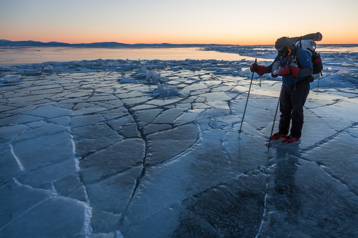 Greg Mills in his ice skates wearing a backpack, standing on cracked ice in Alaska. A beautiful pastel sunset behind him. 