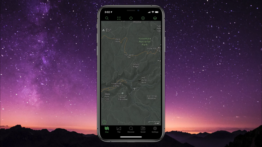A cell phone with the screen displayng a topographical map in Dark Mode, with a dark colored map and light colored writing.  The phone is set against a colorful sunset backdrop.  