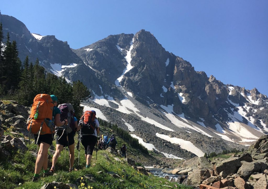 Three backpackers hiking on a trail along side a bubbling creek with a rocky mountain in background.