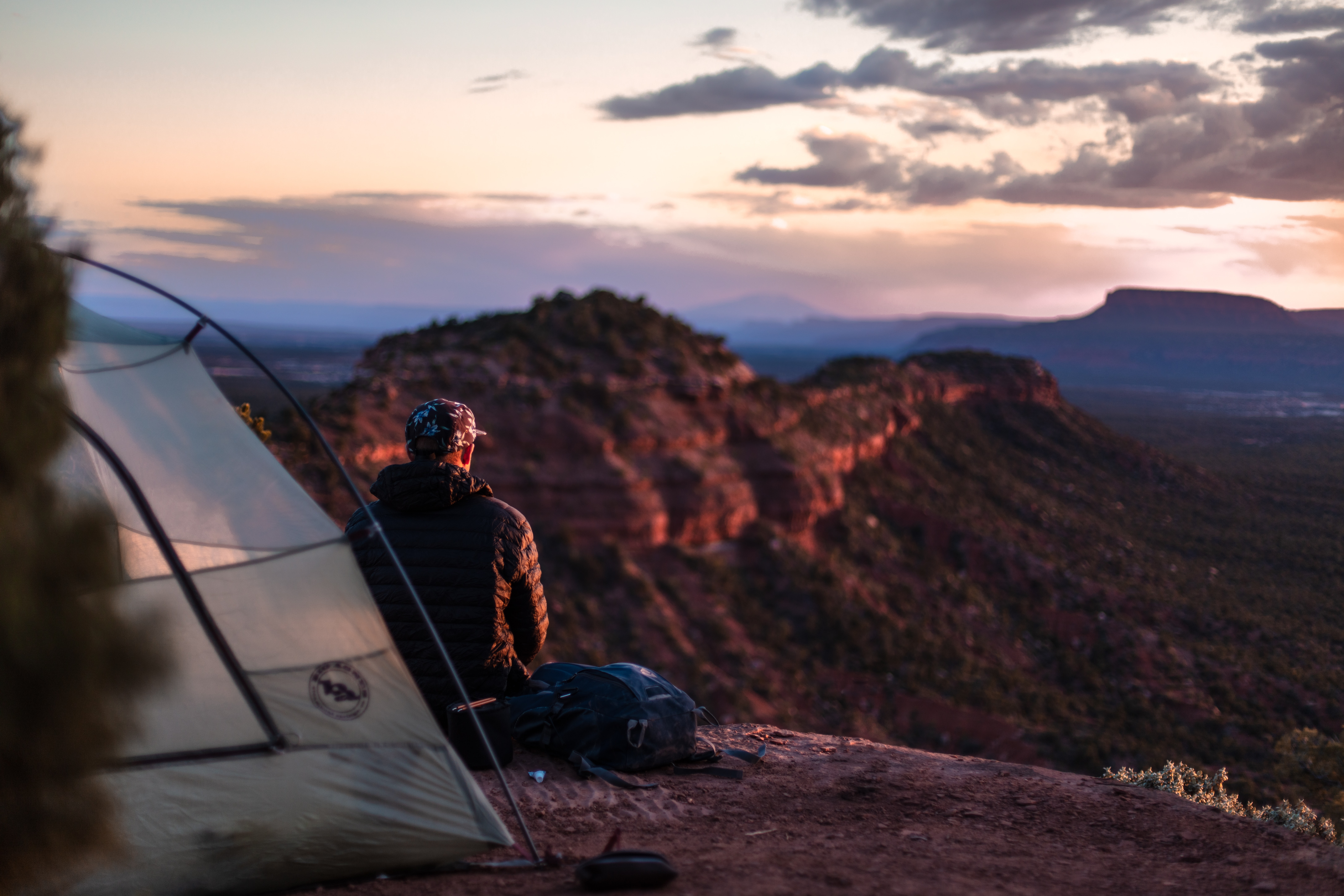 A man sitting on a ledge looking out at a valley, a portion of his tent is in the foreground.  