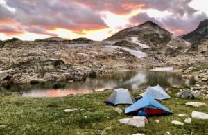 Three backpacking tents along a lake in the mountains with a pink sunset in background.