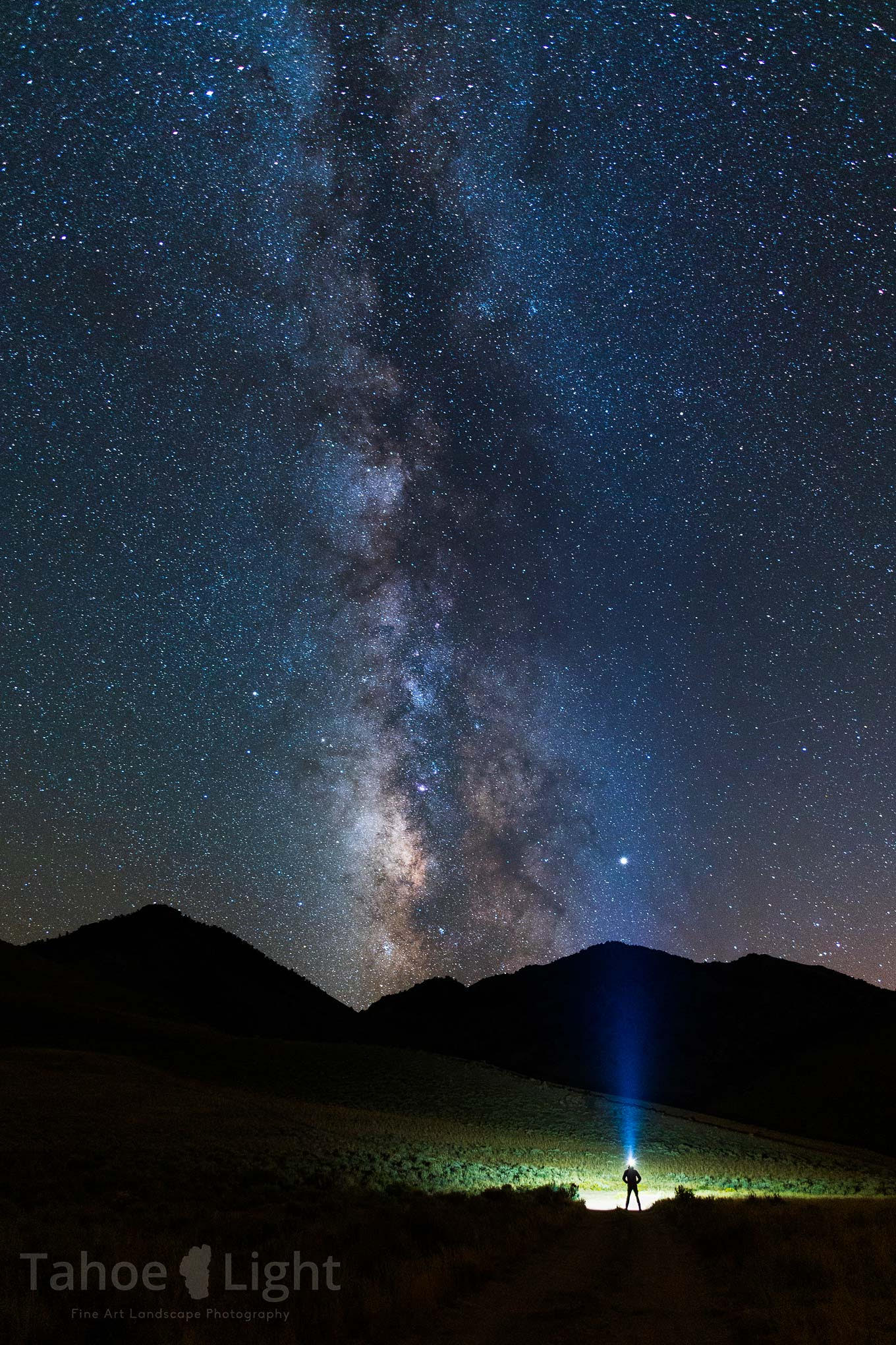 The Milky Way stretches across the night sky with a person with a bright headlamp standing with hands on hips in front of mountain.   