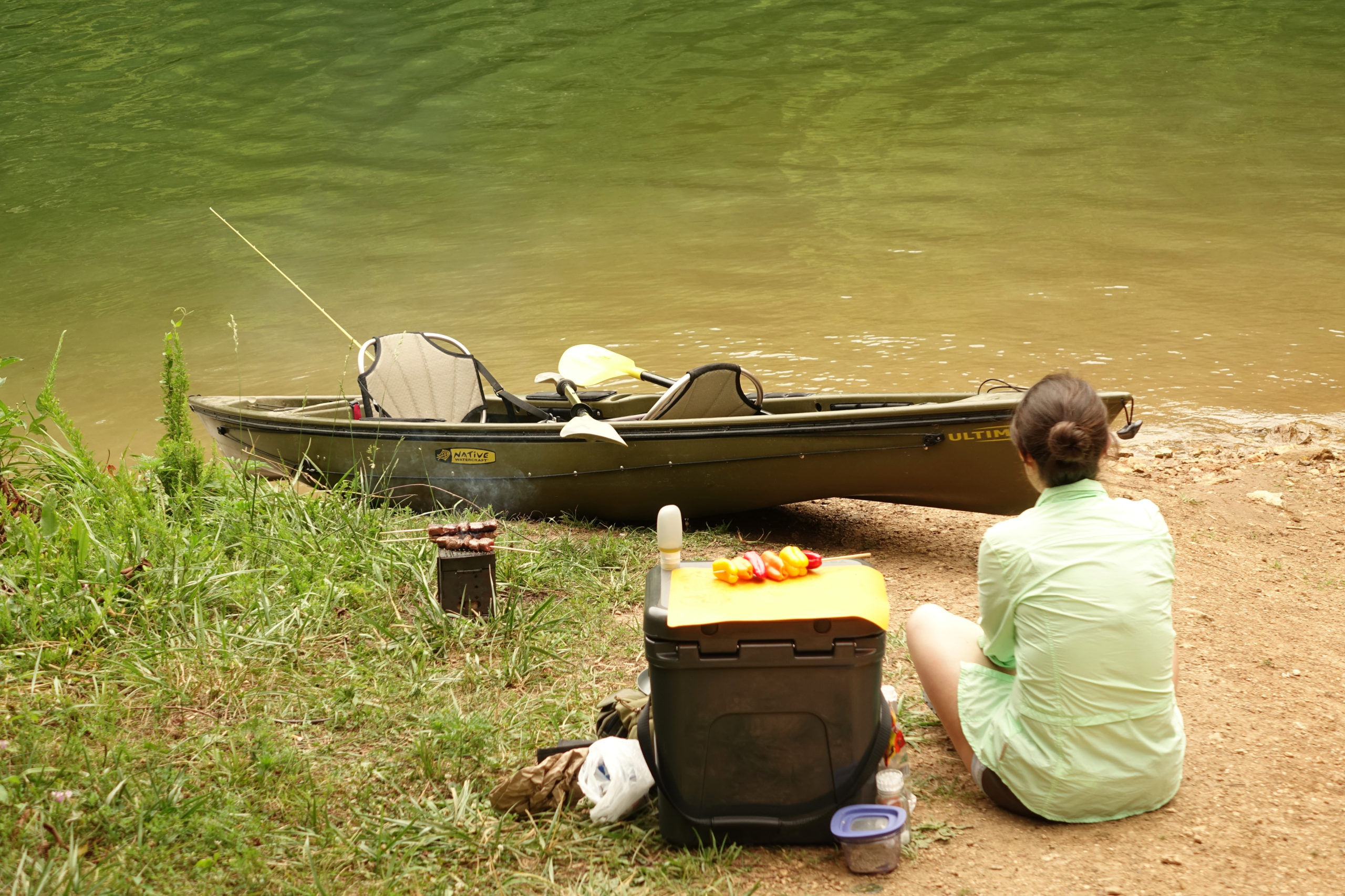 A vegetable kebab sits on a cutting board, which is resting on top of a cooler. A person sits in the sand next to the cooler, gazing at a boat beached on the shore of a lake. 