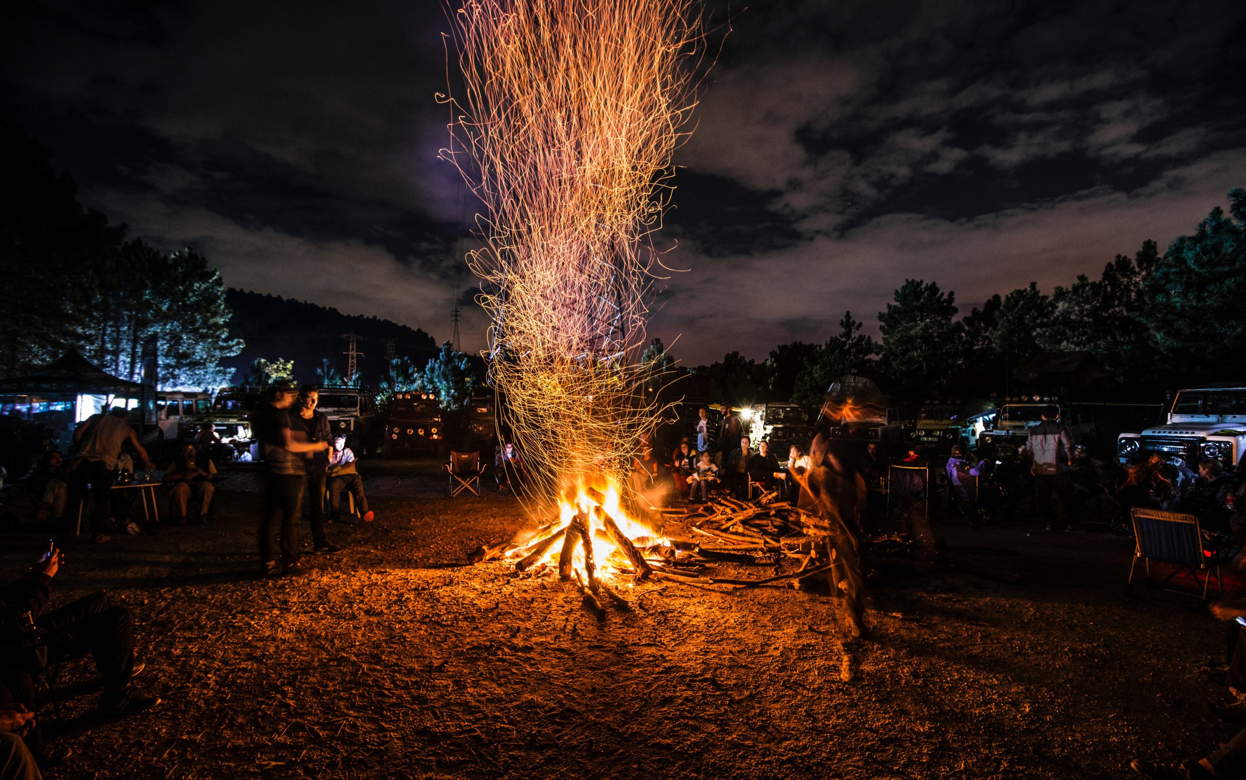 People stand around a bonfire at night with a flock of jeeps parked in the background.