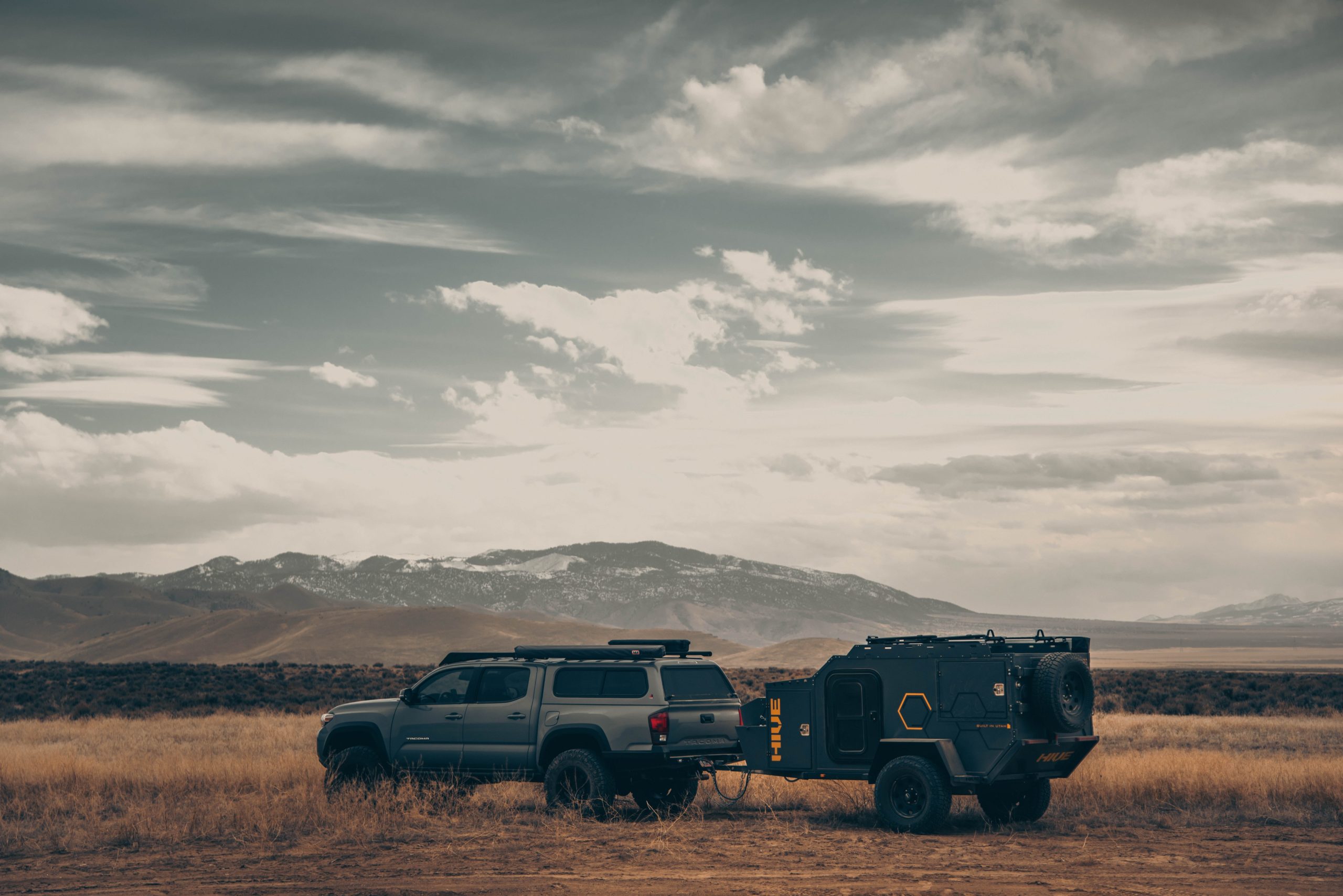 A truck with a covered back and a trailer hitch drives across dry, grassy plains with mountains in the distance.