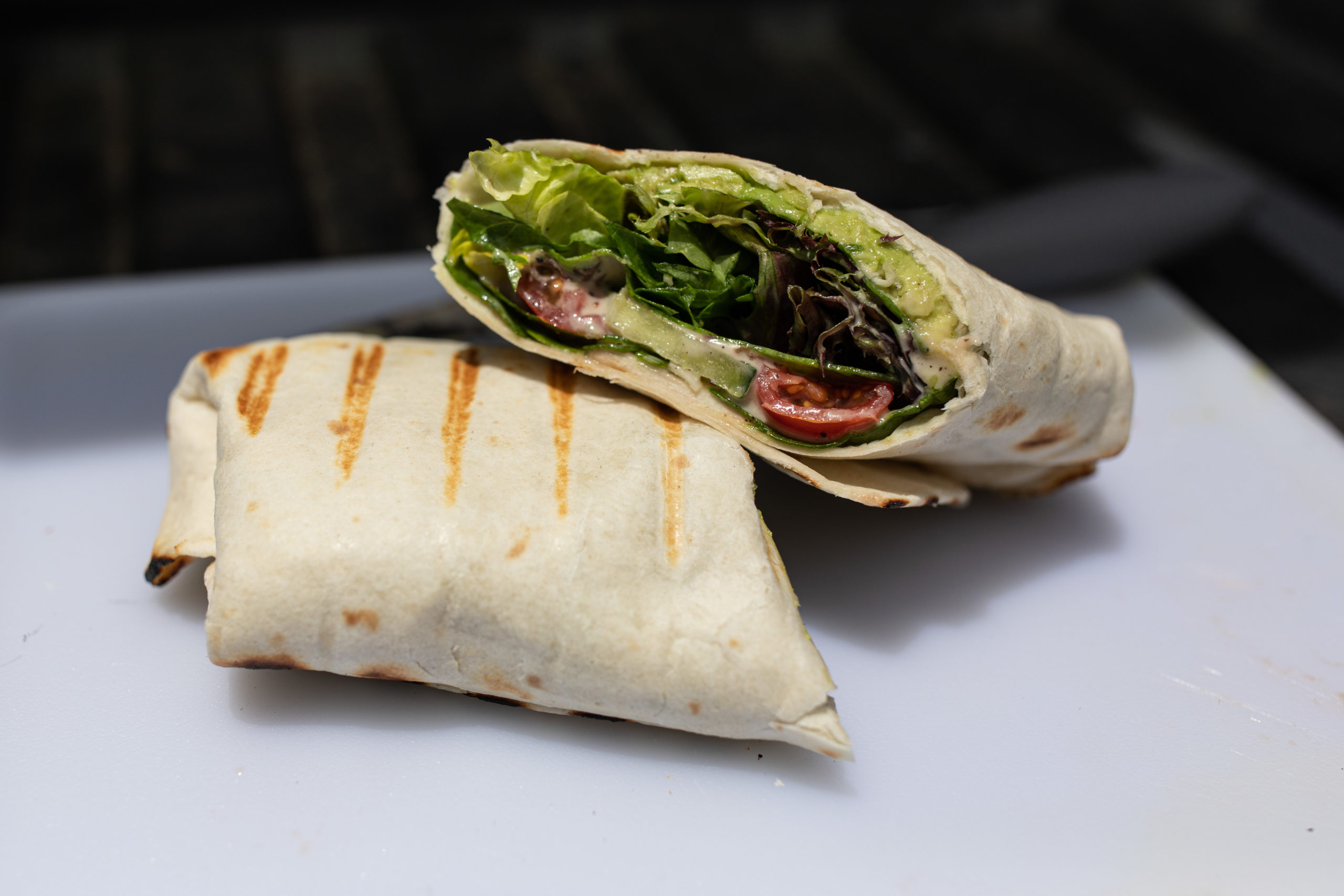 a tortilla wrap filled with avocado, cheese, sliced cucumber, spring mix, halved cherry tomatoes, and dressing sits cut in half on a cutting board.