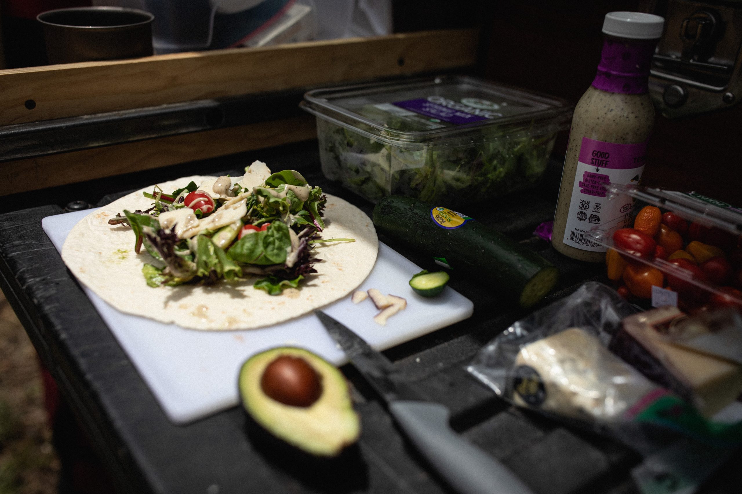 An open tortilla sits on a cutting board, topped with spring mix, halved cherry tomatoes, and salad dressing. A halved avocado, a cucumber, and some cheese also sit on the table.