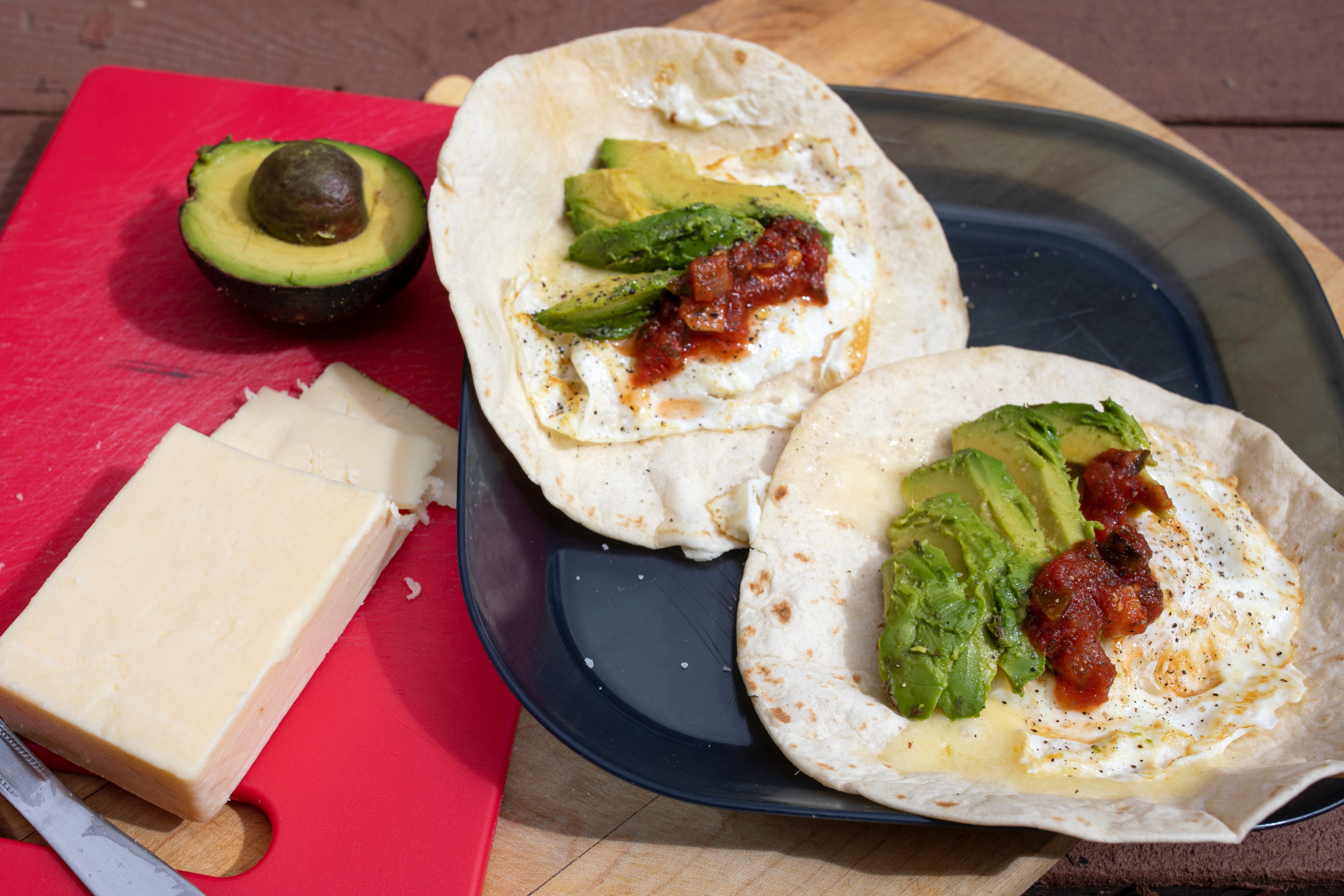 Two tortillas each filled with a friend egg, avocado, salsa, and cheese sit on a plate. A block of cheese and half an avocado sit on a cutting board beside.
