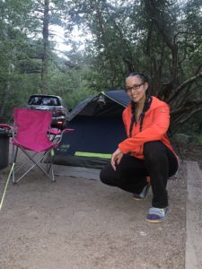 Nicole kneels in a campground next to a camping chair and tent, her car in the background.