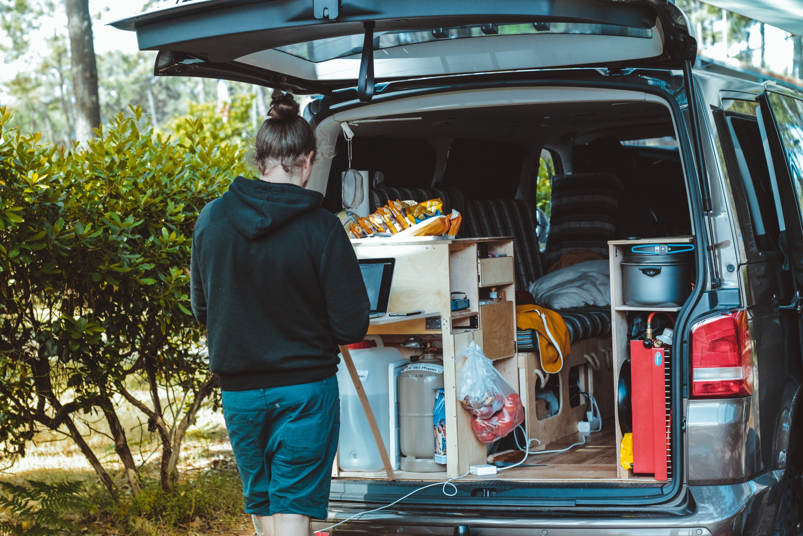 Someone stands with their back to the camera. They work on a laptop placed on a standing desk, which faces out of the back of a built-out van. 