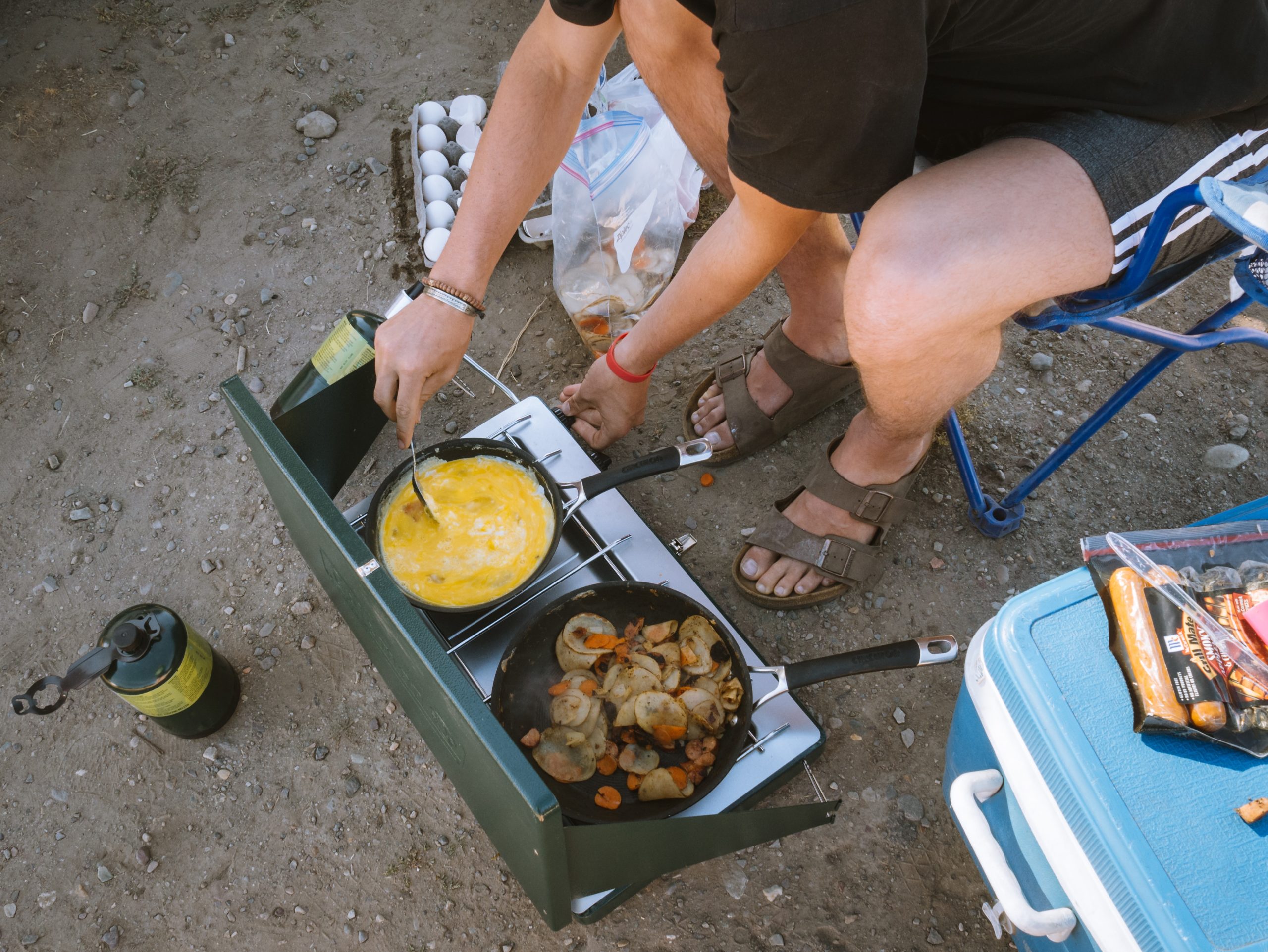 An overlander sits in a camping chair, tending to scrambled eggs in a skillet on a two-burner portable stove. Vegetables sauté in the a pan on the other skillet. A pack of sausages sit on a cooler in the foreground. 