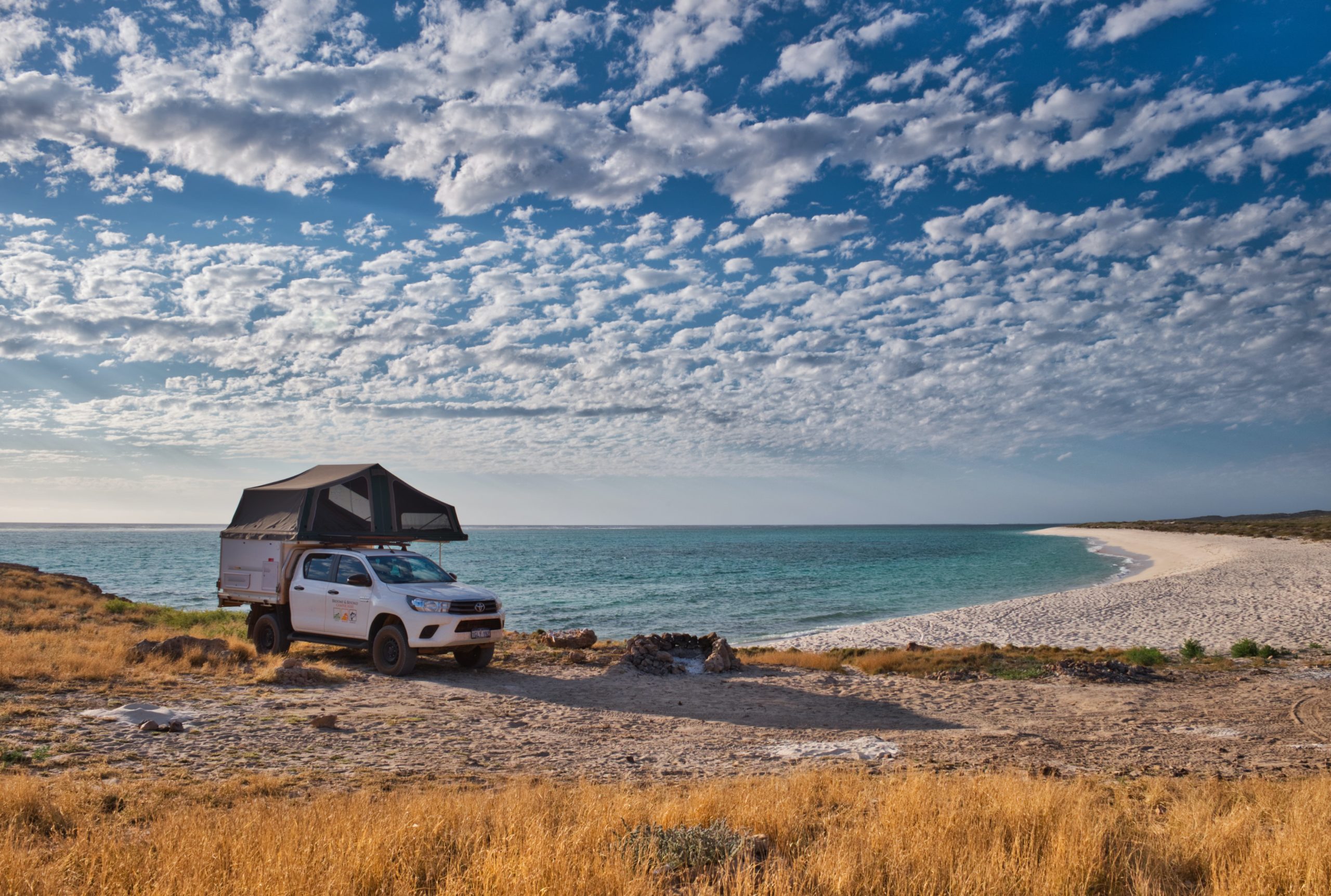 A modified truck with a storage unit in the back and a rooftop tent is parked at a beach on a sunny day. Turquoise water laps behind and stratocumulus clouds cover the blue sky.