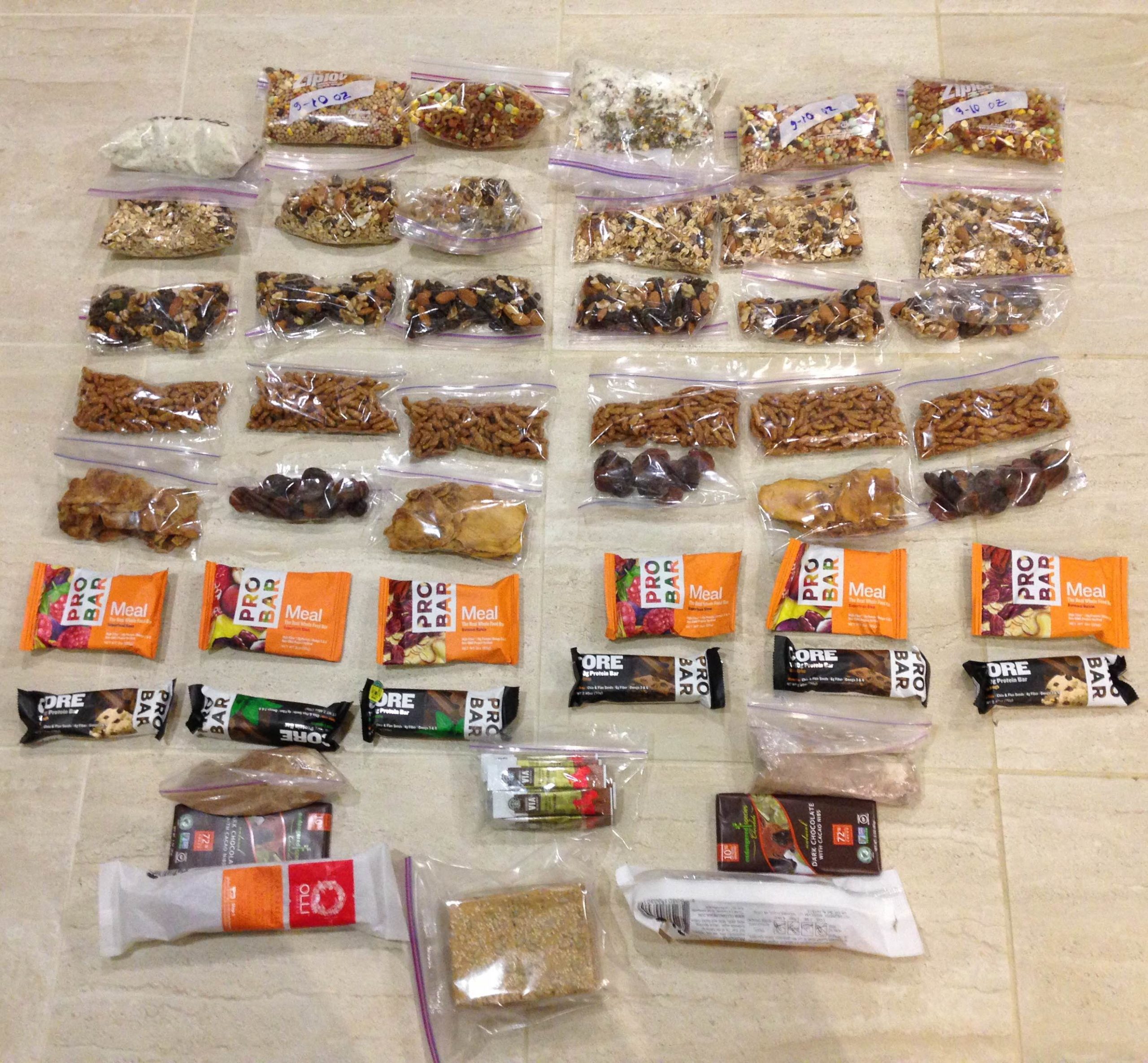 Six days worth backpacking food lies in rows on the floor. Dehydrated meals, trail mix, and dried fruit have been repackaged into small ziplock pages. The allotment also contains bars, instant coffee, and two bars of chocolate.
