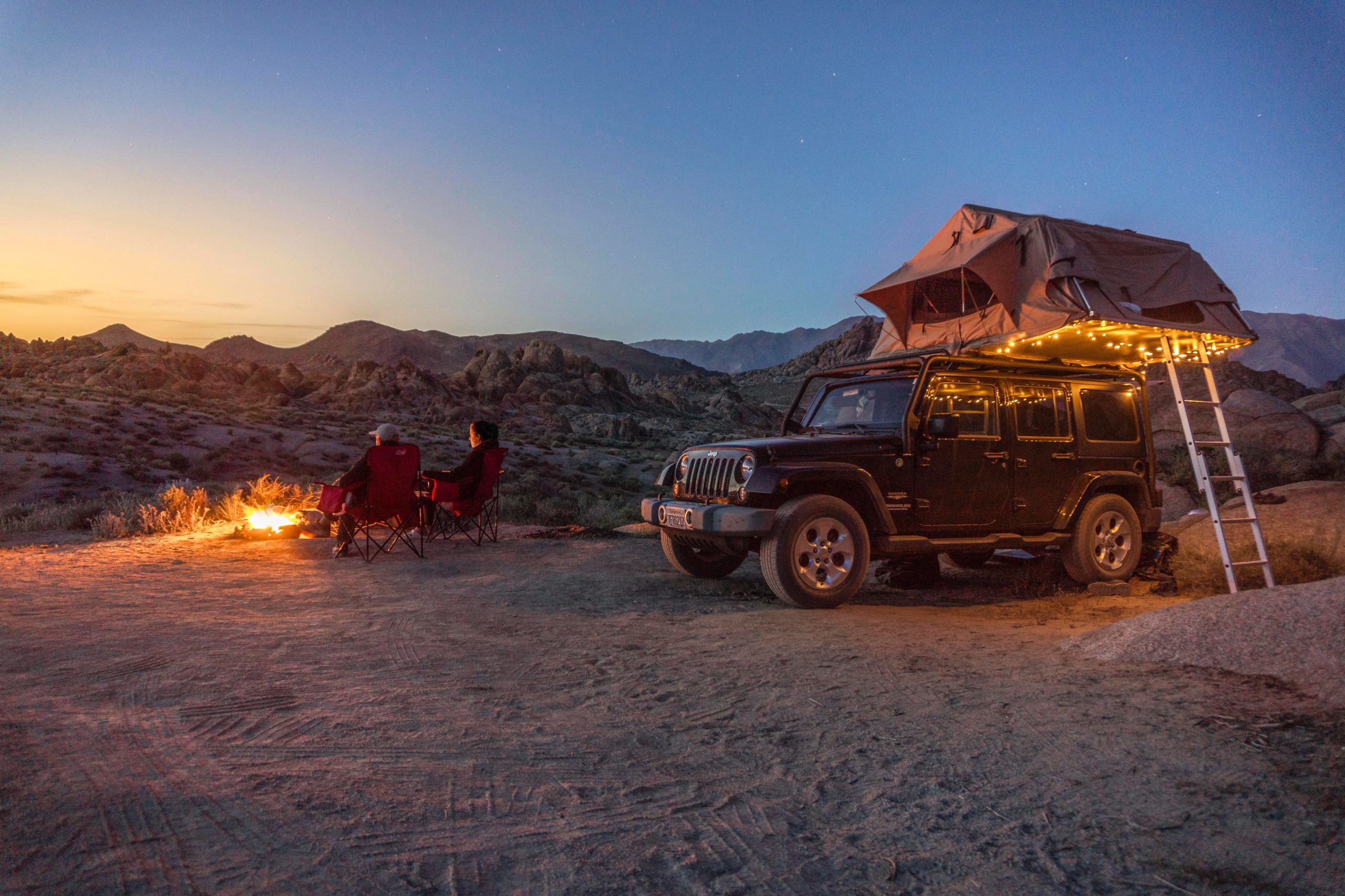 A jeep with a roof tent is parked in the desert. Off to the side, two people sit in camp chairs around a fire, gazing at the sun setting behind the mountains in the distance. 