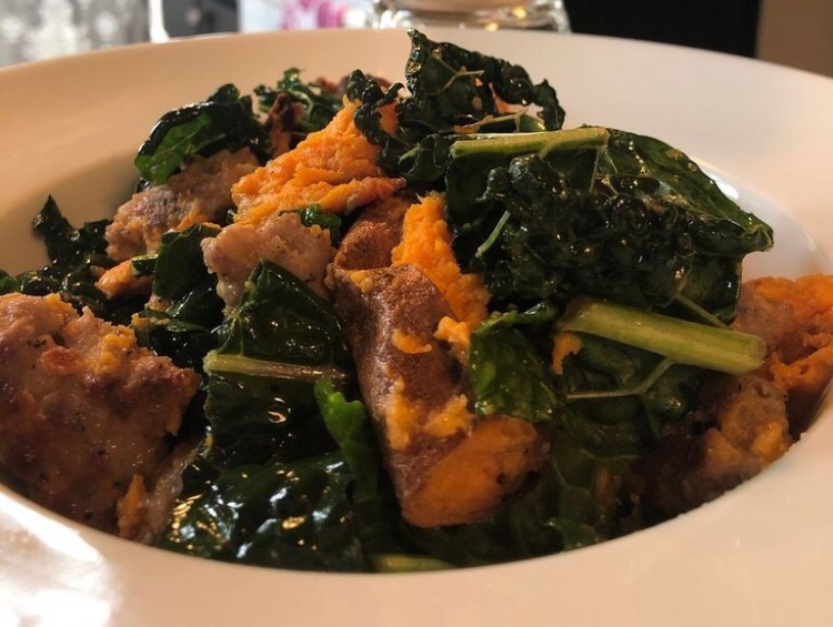 A close up shows a bowl of roasted sweet potatoes, sausage, and kale.