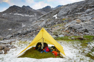 A tent filled with camping gear sits in a snowy alpine meadow surrounded by mountains.