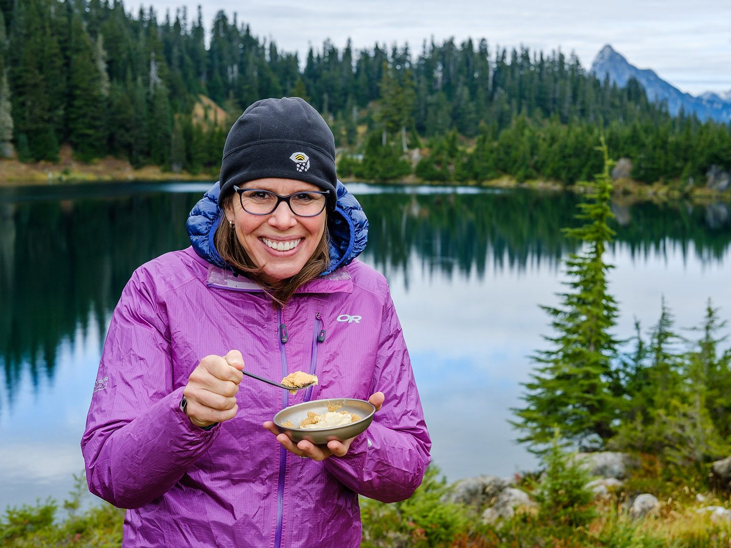 Aaron Owens Mayhew smiles while eating a bowl of pudding in the backcountry. An alpine lake and mountains are in the background.