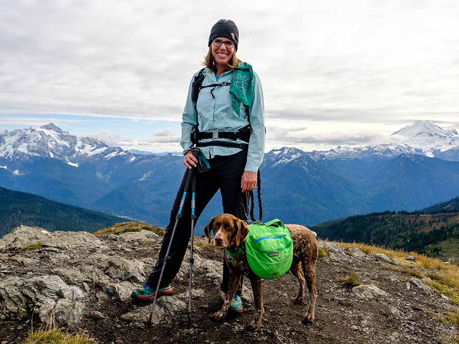 Aaron poses for the camera with her dog, who's loaded down with a dog backpack. Aaron holds her hiking poles and wears a backpack. They are standing in the alpine, with snowcapped-mountains behind.