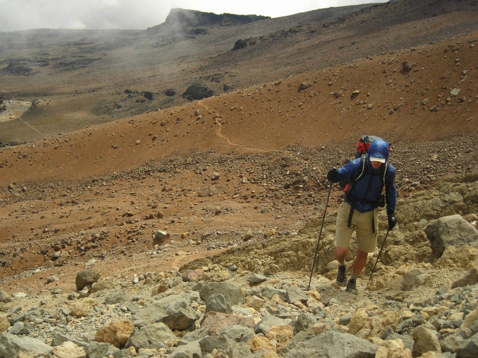 Lichter backpacks up a barren climb in Africa. He wears a jacket, gloves, and a brimmed hat.