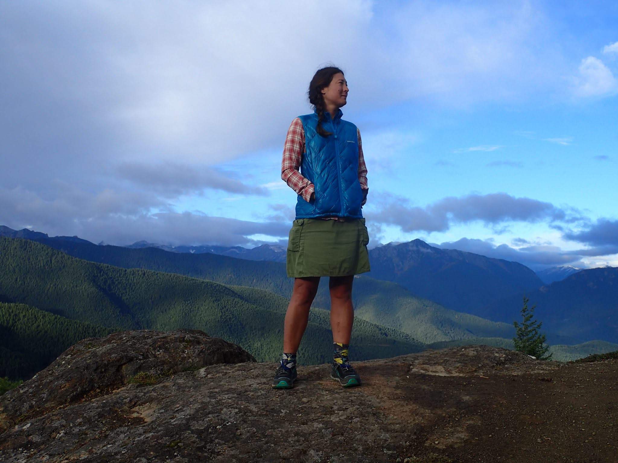 Snorkel stands on a mountain top, looking off her shoulder into the distance.  Green and blue mountains extend behind her.