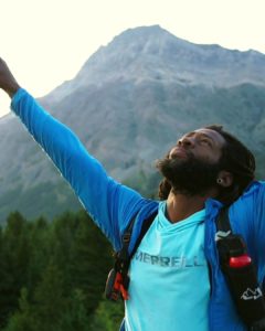 Will Robinson raises his arms above his head. His eyes look up to the sky. A mountain looms behind him.