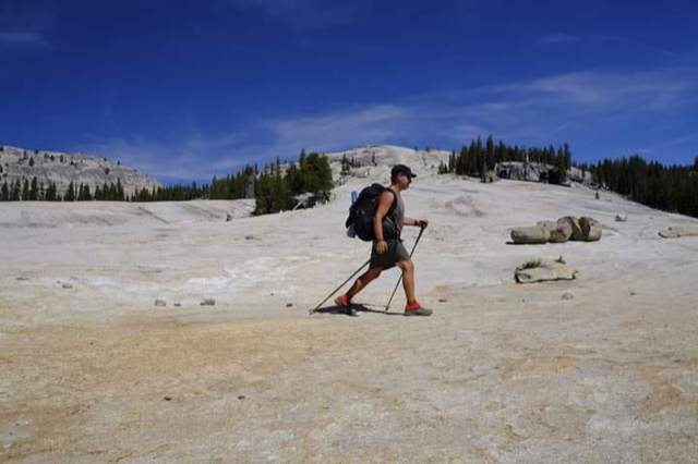 Rue McKenrick hikes across a barren desertscape. He's carrying hiking poles and a backpacking pack.