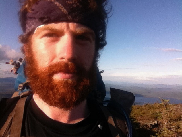 Badger poses for a stoic selfie during his AT thru-hike. He's wearing a headband and sports a thick red beard. Blue-green mountains extend behind him.