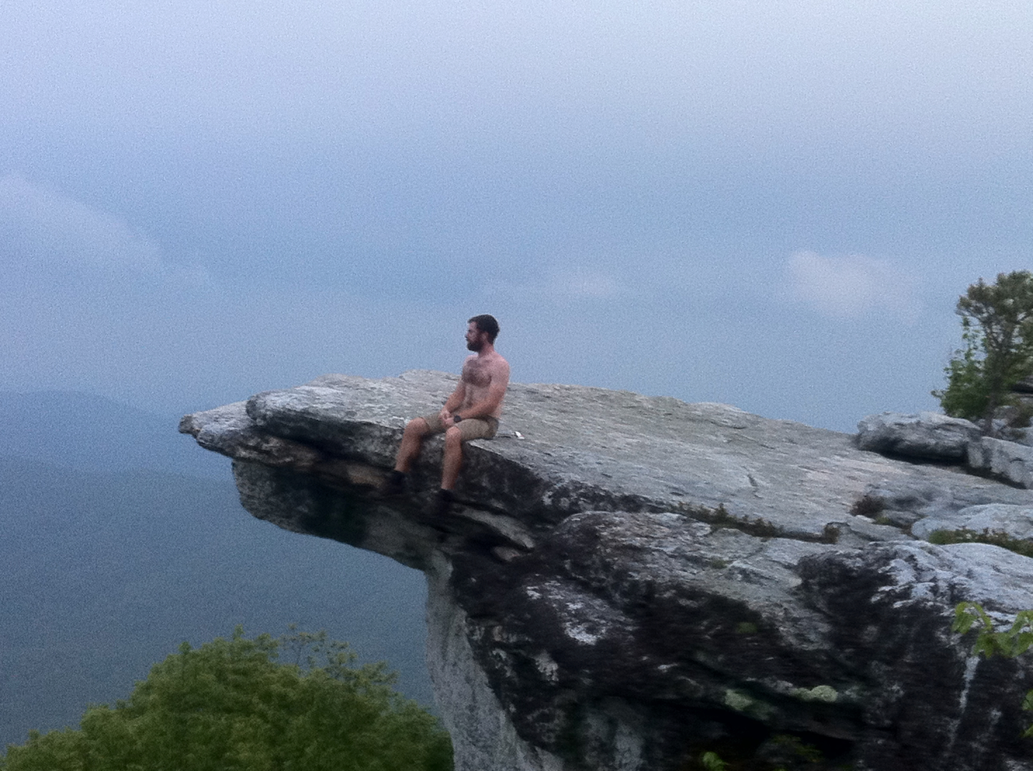 Badger sits on the rocky ledge of McAfee Knob. He's shirtless and sis feet are dangling off the side into the open air below. 