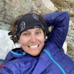 Mary smiles while lying down and resting her head on a rock. She's wearing a purple jacket, gloves, and a black buff around her ears.