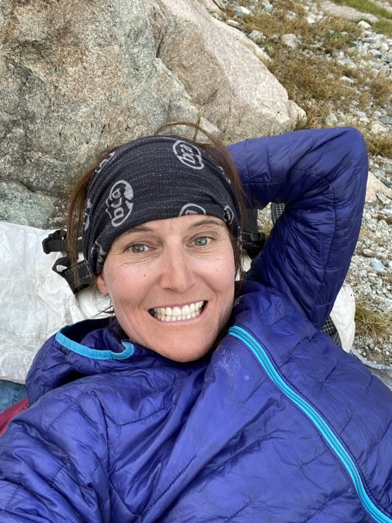 Mary smiles while lying down and resting her head on a rock. She's wearing a purple jacket, gloves, and a black buff around her ears.