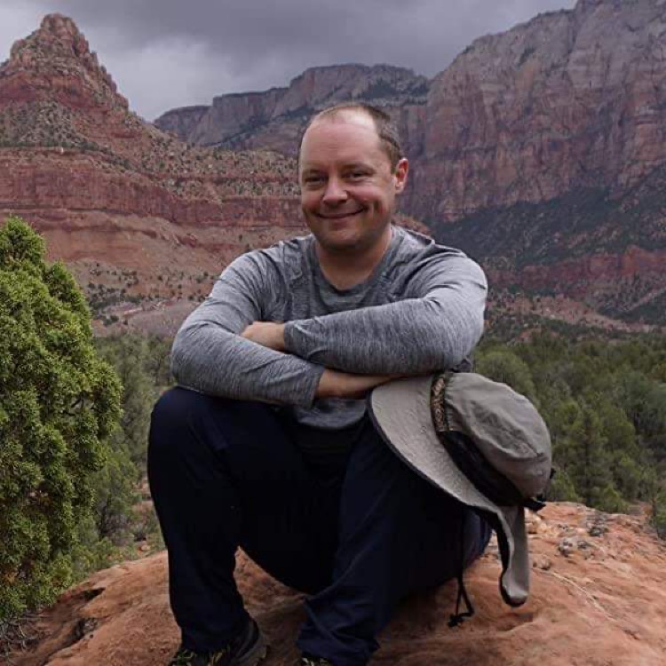 Scott Turner smiles for the camera while sitting on a rock with his arms crossed on his knees. Red and gold canyons loom in the background. 