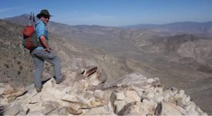 Scott Turner smiles over his shoulder while standing on a rocky overlook. A desert valley with surrounding mountains sprawls out behind him. He's loaded down with his hiking backpack and apparel.