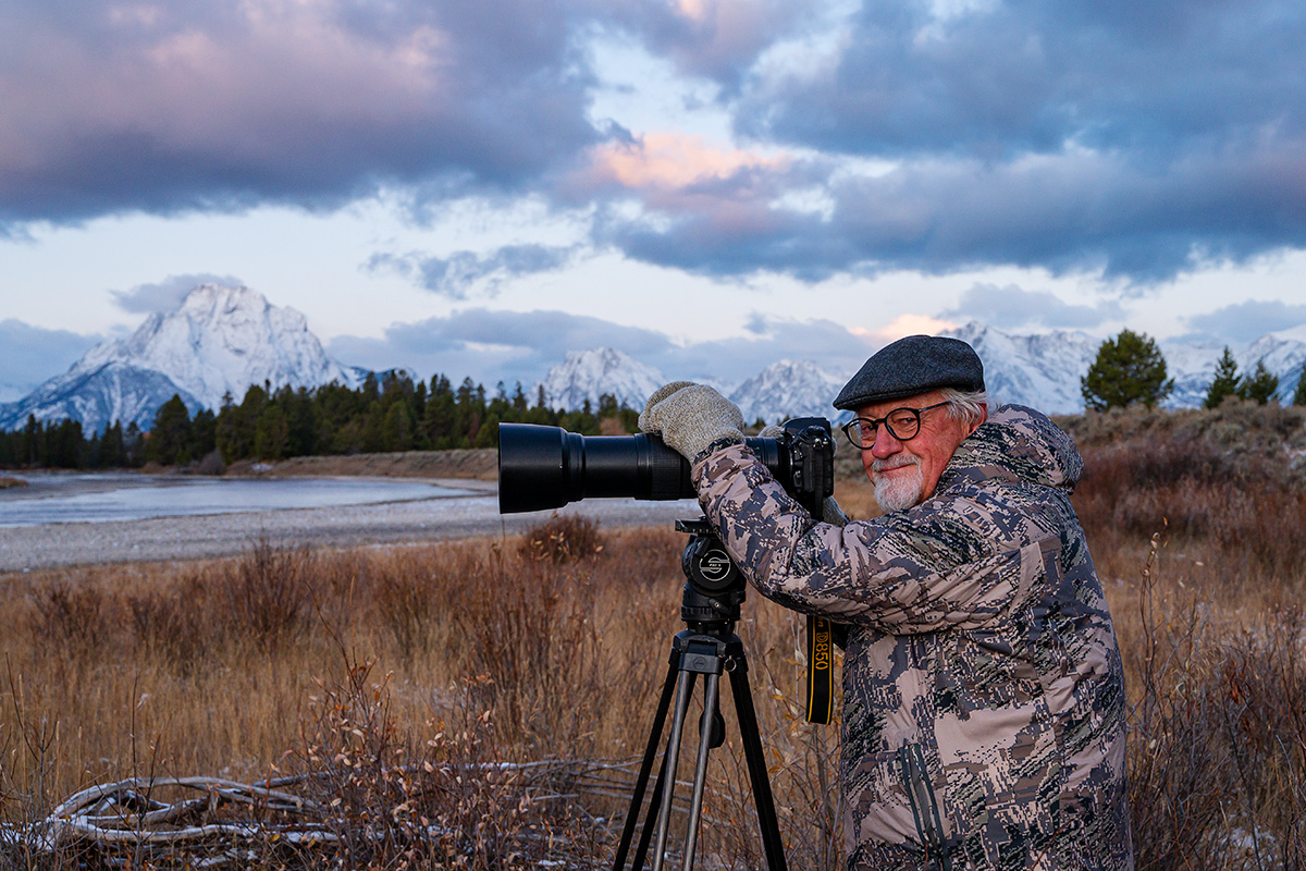 Tom Mangelsen looks towards the camera as he sits in a field with his long-lens camera set up on a tripod. Snowy Tetons loom into the cloudy sky in the distance.