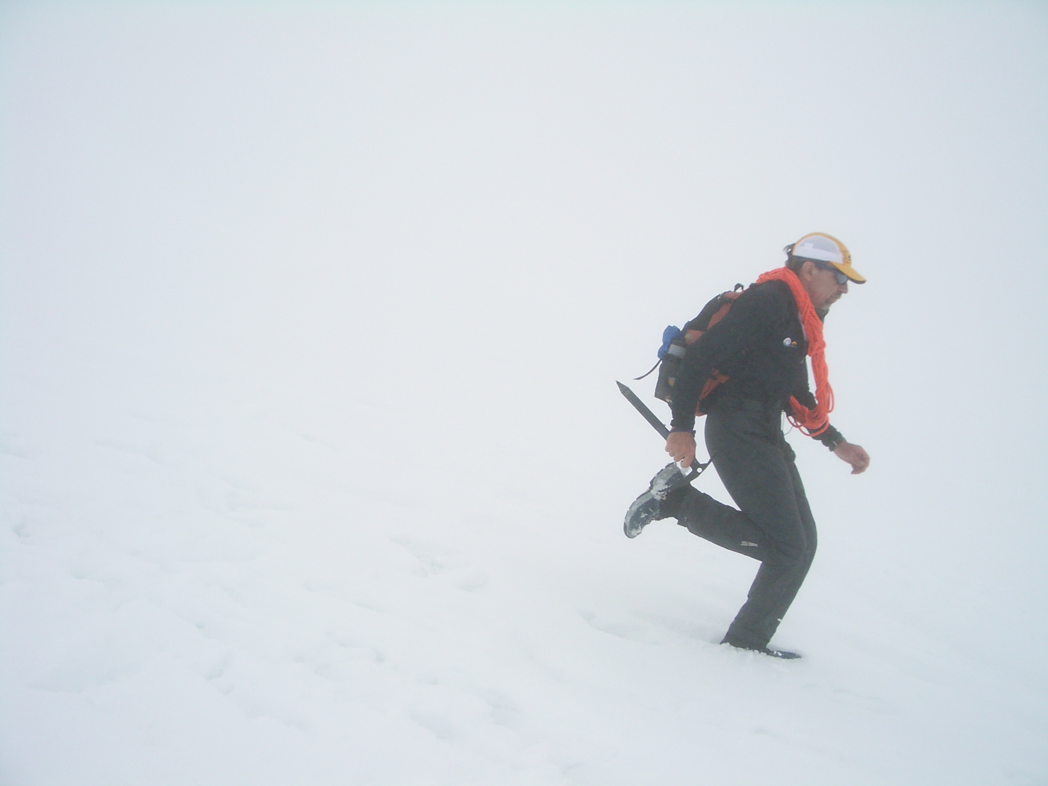 Buzz runs down a snow slope in a white-out. He's carrying an ice ax, shouldering a cord of rope, and wearing a backpack.
