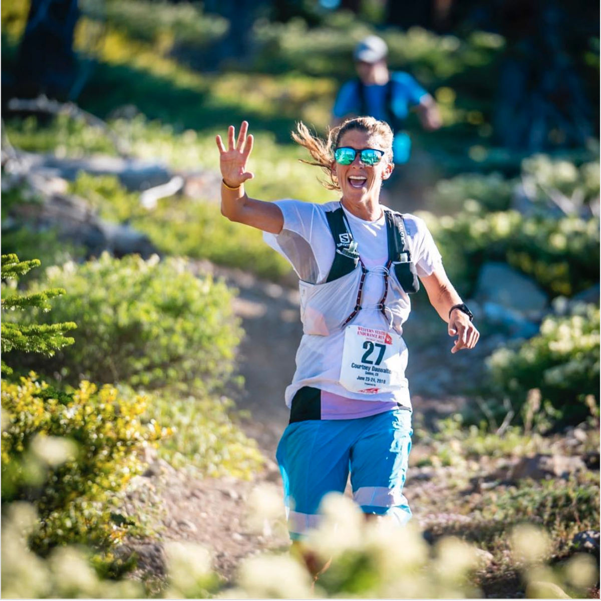Courtney smiles and waves as she runs down a lush trail near Lake Tahoe during the Western States 100 Endurance Run. She's wearing long blue shorts, a white t shirt, reflective sunglasses, and a hydration pack.