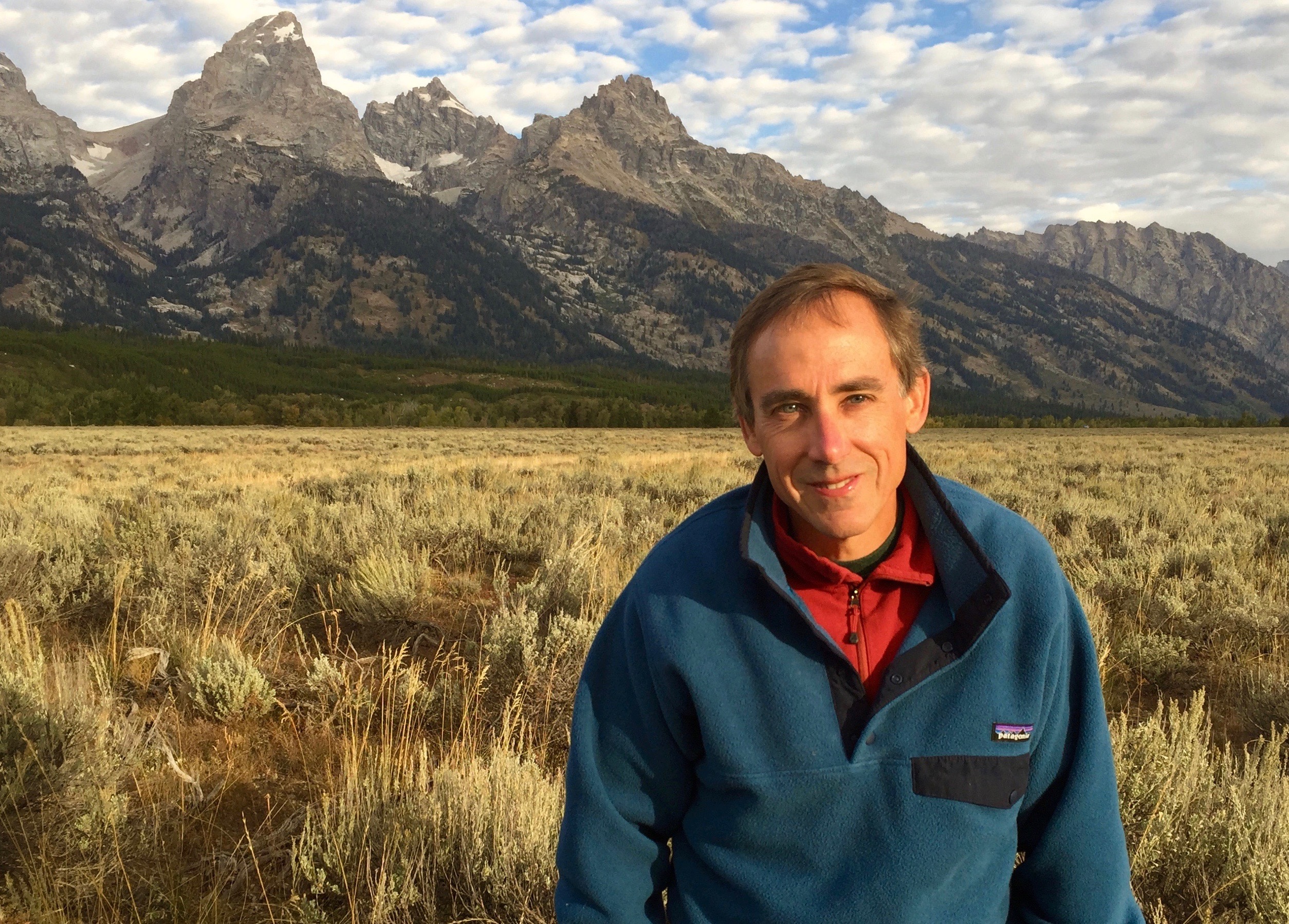 Todd Wilkinson smiles for the camera while sitting in a sunny field at the base of the Tetons.
