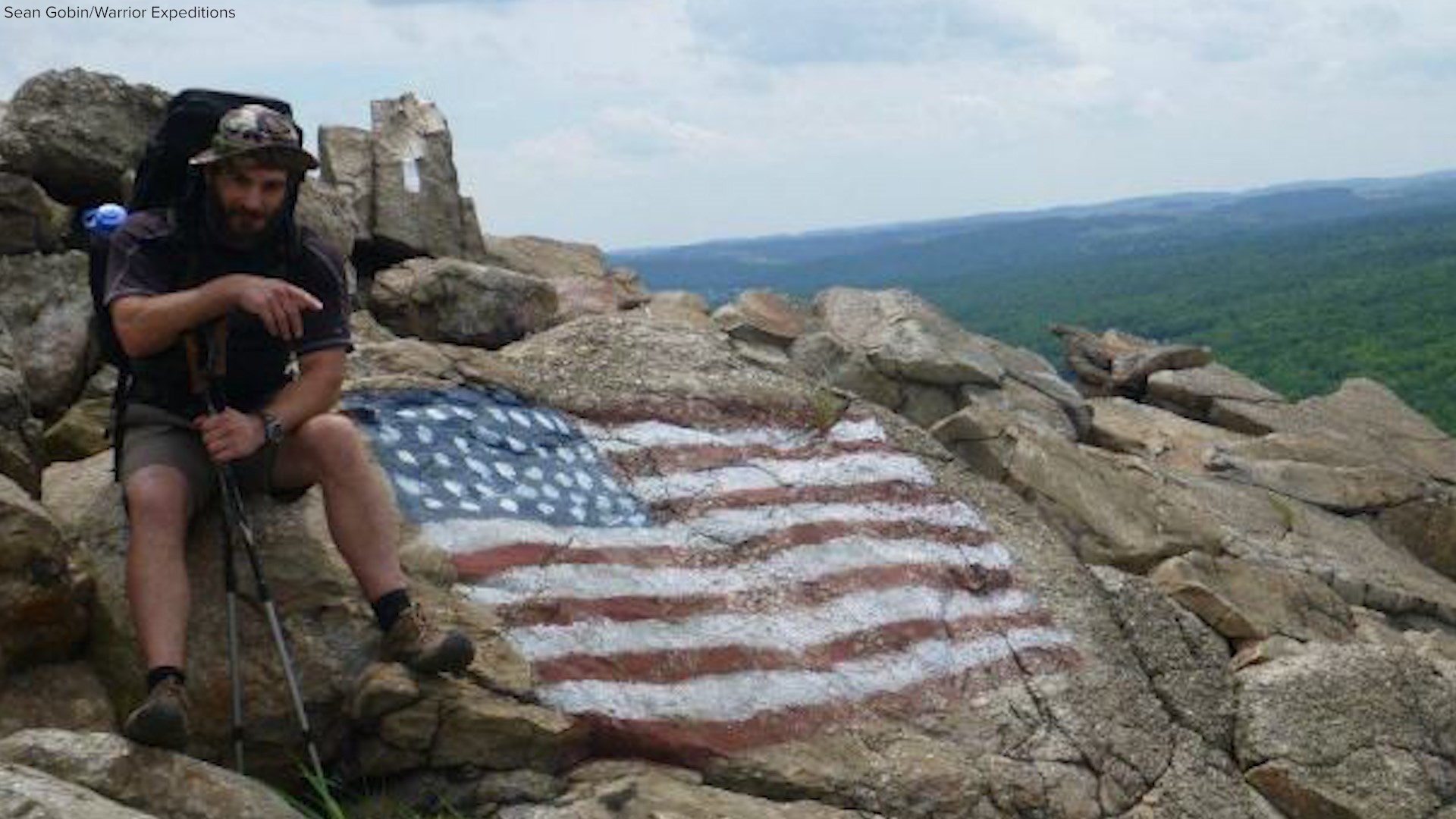 Sean Gobin sits on a rock painted with an American Flag. He's wearing his backpacking pack and holding poles. 