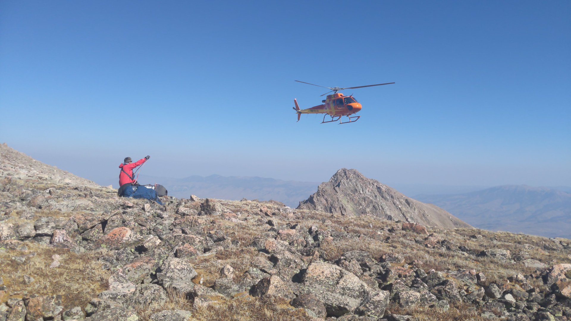 A search and rescue person sits on a rocky alpine field. A helicopter circles overhead. 