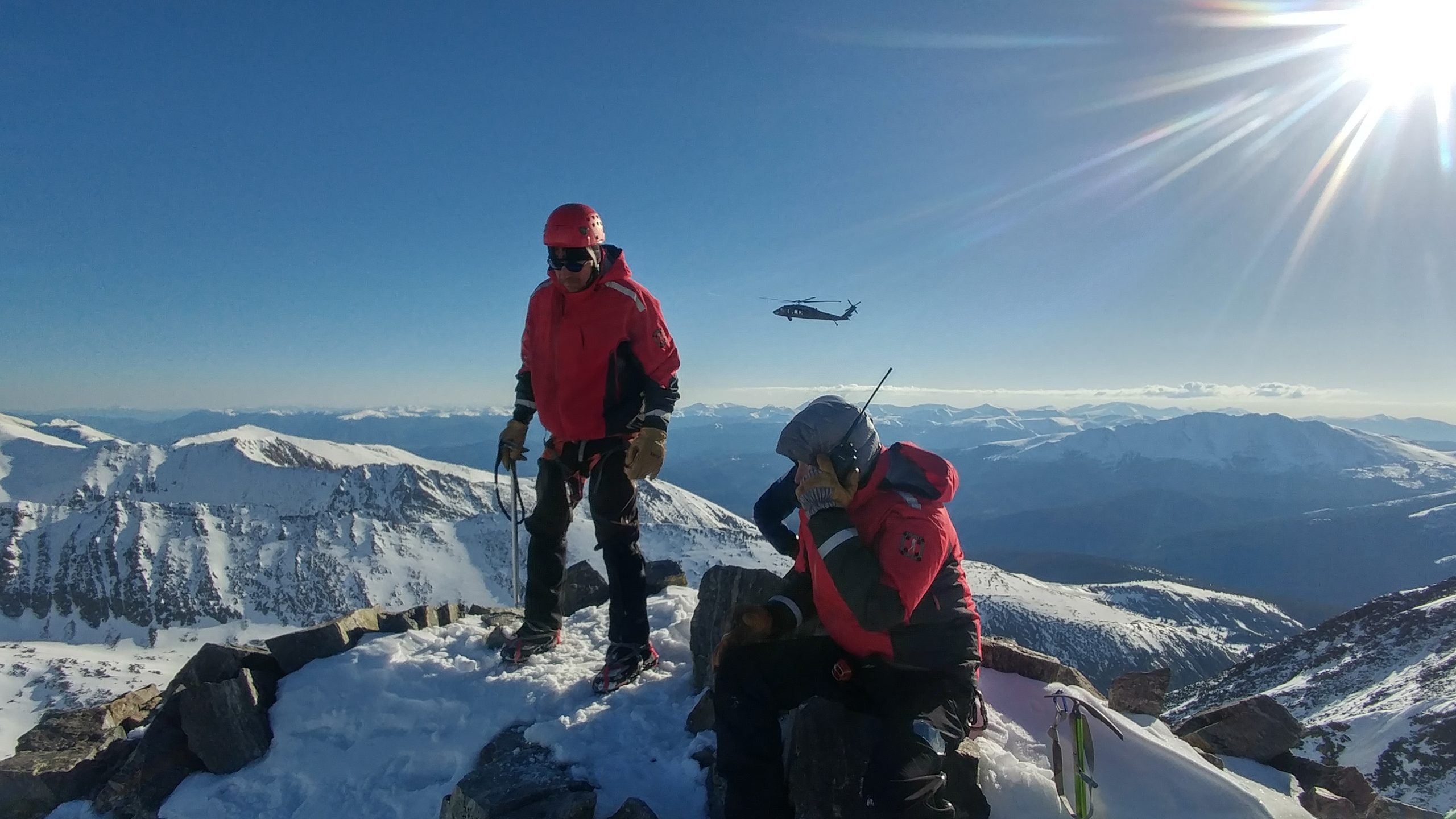 Two people are on top of a snowy mountain. One sits while holding a walkie talkie up to his ear. The other stands with a helmet on his hand, rope in his band, and crampons on his foots. A helicopter flies over mountains in the distance.