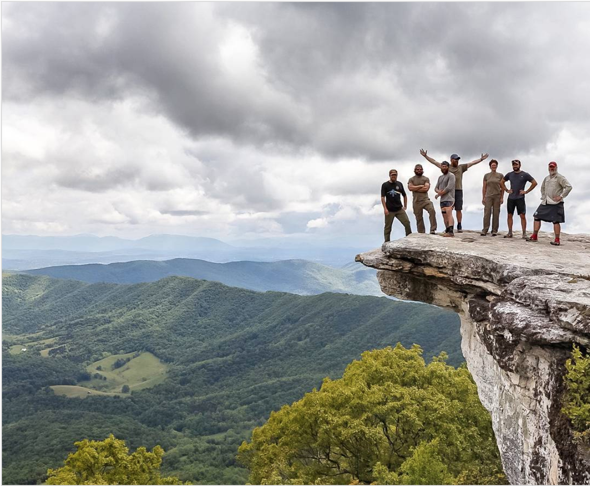 A group of Warrior Expeditions Vets stand on top of McAfee Knob. Green mountains ripple into the distance under a cloudy sky.