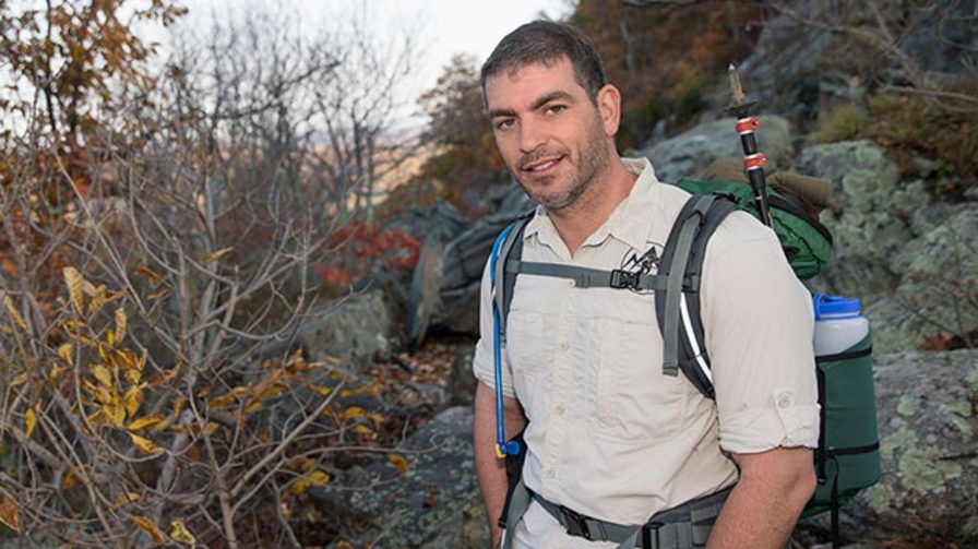 Sean Gobin smiles while standing on a rocky trail. He's wearing a backpack and button-down shorts sleeve shirt. 