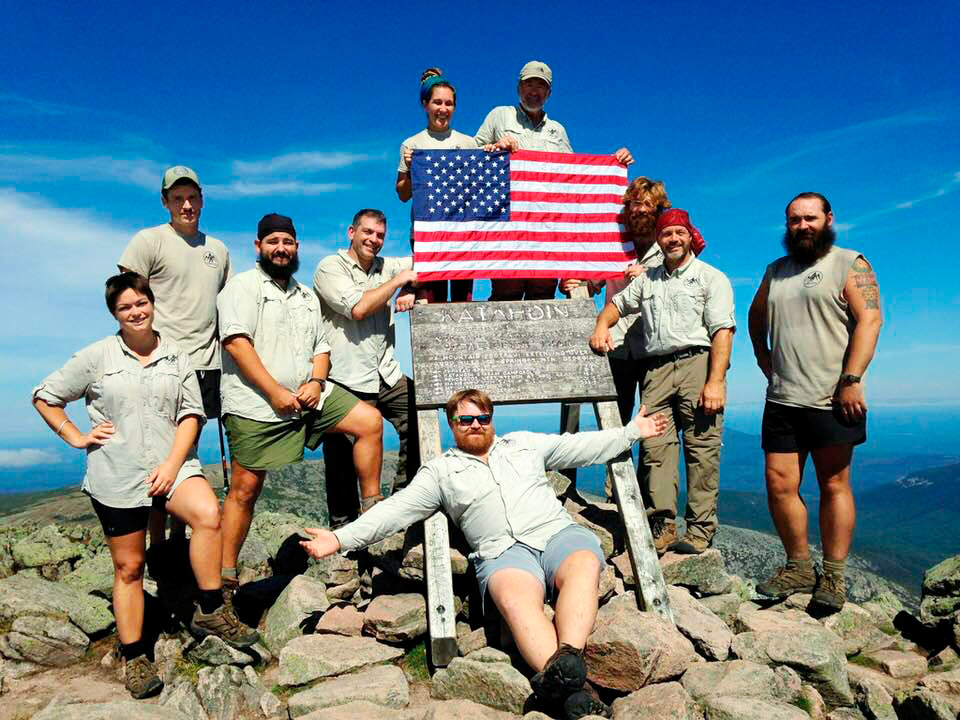 A Warrior Expeditions group stands on top of Mount Katahdin. They are posing and smiling around the sign, and two people are holding an American flag.