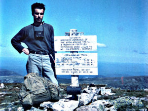 Earl Shaffer stands next to the sign on top of Mount Katahdin. He wears binoculars, a long sleeve tee and jeans. His pack sits on a rock in front of him.