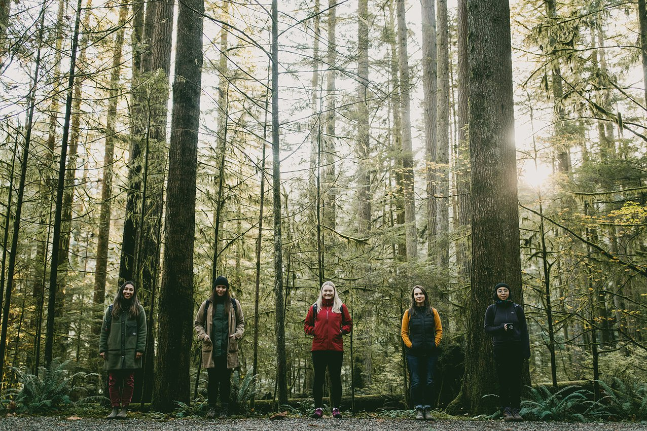 Five people stand in a line in a forest and smile for the camera.